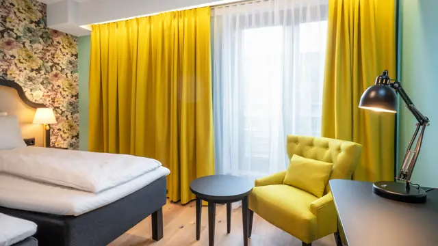 To enkeltsenger, blomstret tapet bak sengene, gul stol, arbeidspult og store vinduer med store gule gardiner på twin room på Thon Hotel Cecil i Oslo sentrum