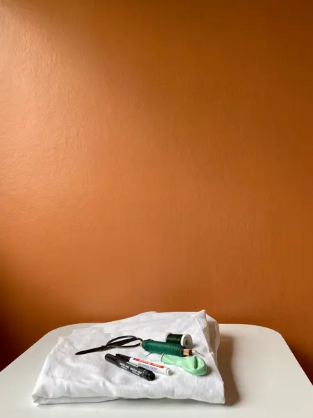 Hvit laken, tusj, tråd, balong og saks som ligger på et bord med oransje bakgrunn