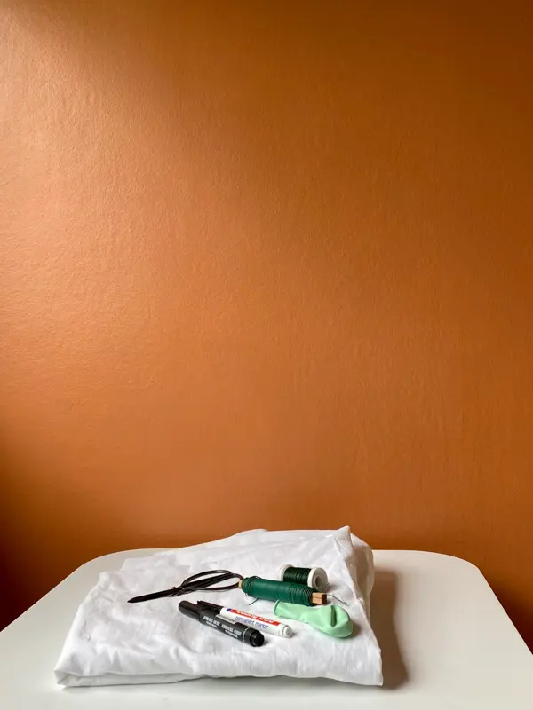 Hvit laken, tusj, tråd, balong og saks som ligger på et bord med oransje bakgrunn