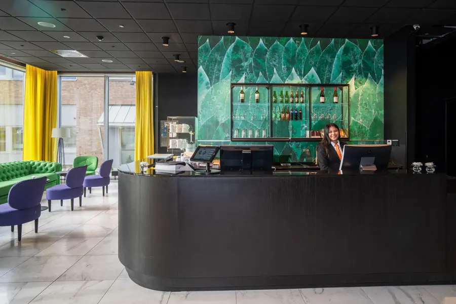 Lyst gulv, svart tak, grønn sofa, lilla stoler, vindu fra gulv til tak, gule gardiner, svart resepsjonsdisk, resepsjonist, grønn tapetvegg. 