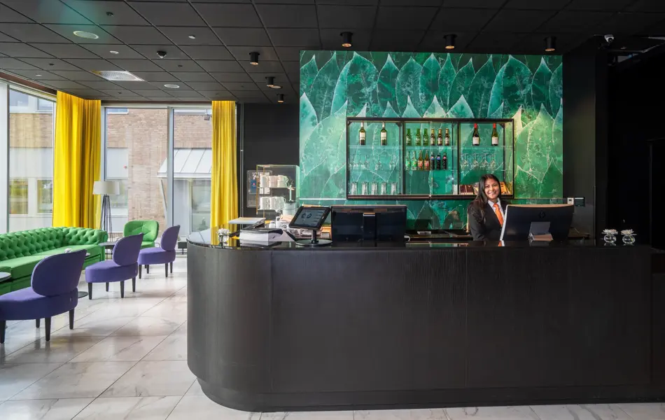 Lyst gulv, svart tak, grønn sofa, lilla stoler, vindu fra gulv til tak, gule gardiner, svart resepsjonsdisk, resepsjonist, grønn tapetvegg. 