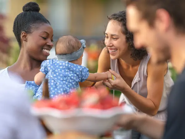 A young African American mom holds her adorable young daughter in her lap at an outdoor dining table. The girl is smiling and reaching toward mom's friend, who's smiling at her.