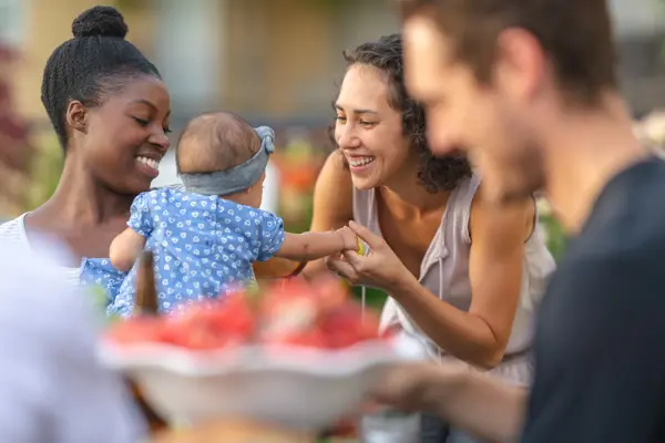 A young African American mom holds her adorable young daughter in her lap at an outdoor dining table. The girl is smiling and reaching toward mom's friend, who's smiling at her.
