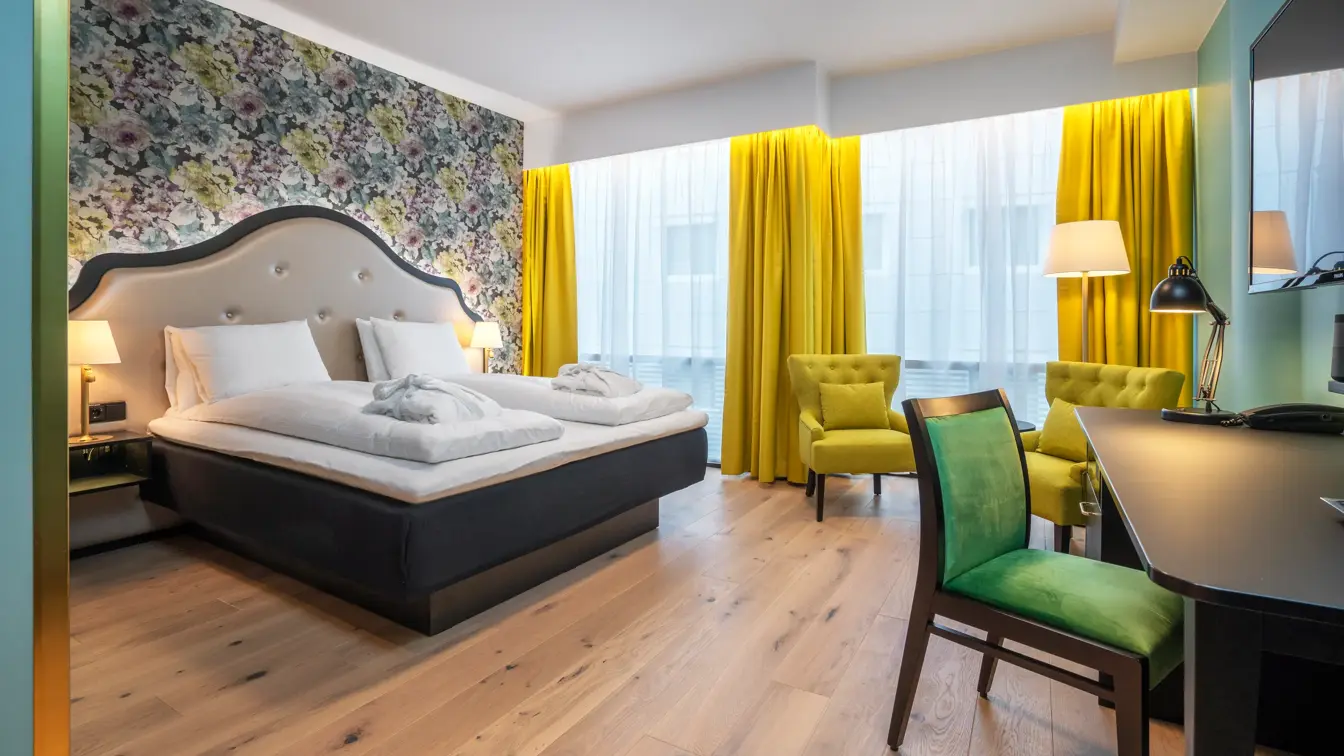 Dobbeltseng med morgenkåper. Skrivebord med grønn stol. Sittegruppe med gule stoler. Blomstrete tapeter, gule gardiner og store vinder i superior rom på Thon Hotel Cecil