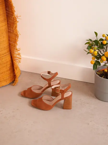 Brune semsket sandaler med hæl og åpen tå, med appelsinplante og oransje teppe i bakgrunnen.