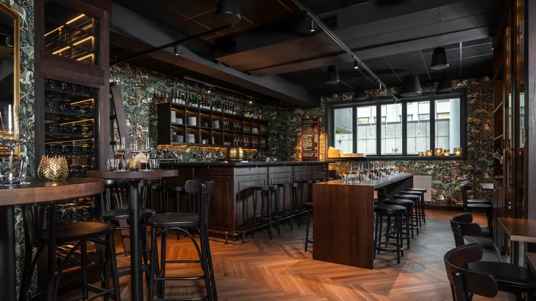 Grotten øl- og vinbar med bardisk med tilhørende runde barkrakker, mørk tapet med tegning av trær, hester og andre dyr og planter, langbord med tilhørende mørke krakker, , høye barbord stort vindu, parkettgulv.