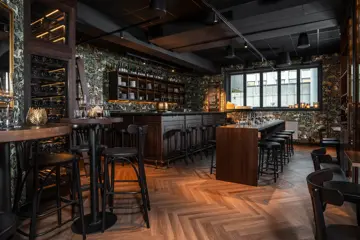 Grotten øl- og vinbar med bardisk med tilhørende runde barkrakker, mørk tapet med tegning av trær, hester og andre dyr og planter, langbord med tilhørende mørke krakker, , høye barbord stort vindu, parkettgulv.