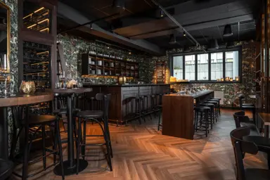 Grotten øl- og vinbar med bardisk med tilhørende runde barkrakker, mørk tapet med tegning av trær, hester og andre dyr og planter, langbord med tilhørende mørke krakker, , høye barbord stort vindu, parkettgulv.