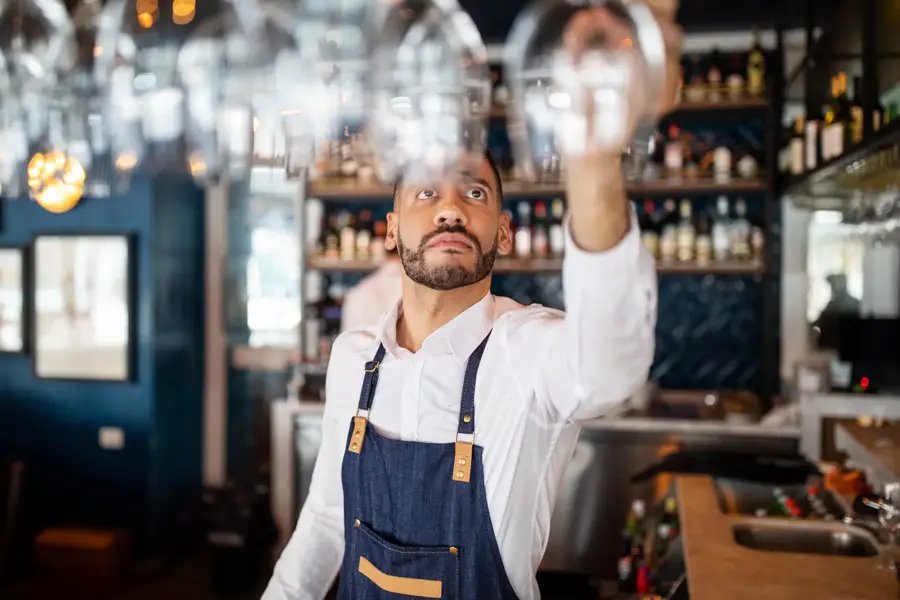 Mixed race barman working at the cafe. Bartender taking a wineglasses from the overhead rack at bar counter.