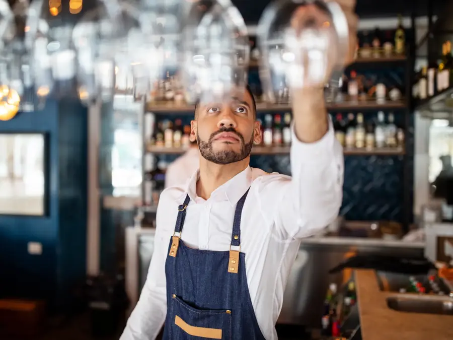 Mixed race barman working at the cafe. Bartender taking a wineglasses from the overhead rack at bar counter.