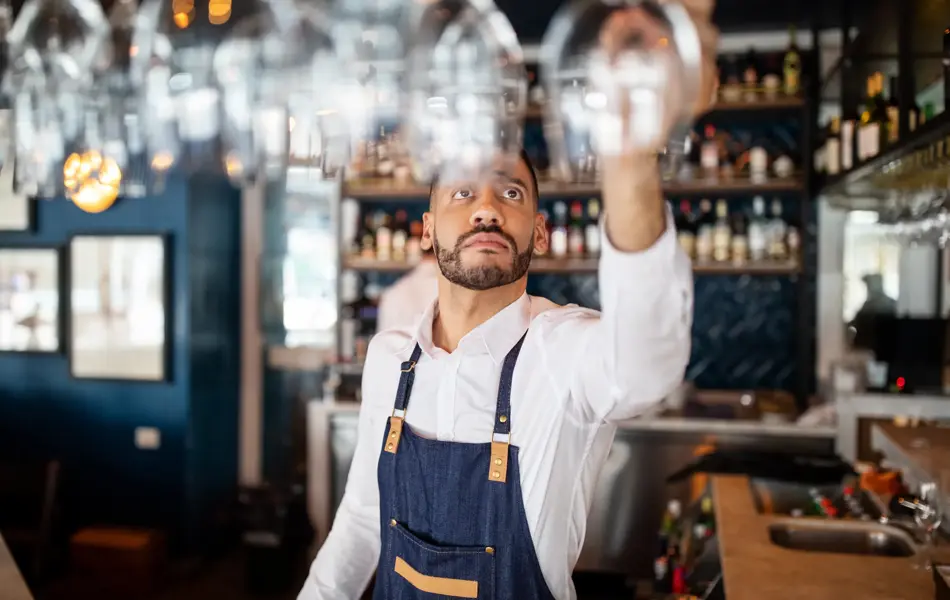 Mixed race barman working at the cafe. Bartender taking a wineglasses from the overhead rack at bar counter.