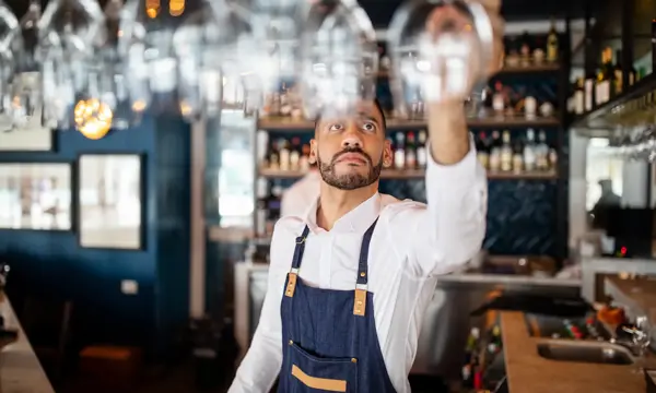 Mixed race barman working at the cafe. Bartender taking a wineglasses from the overhead rack at bar counter.