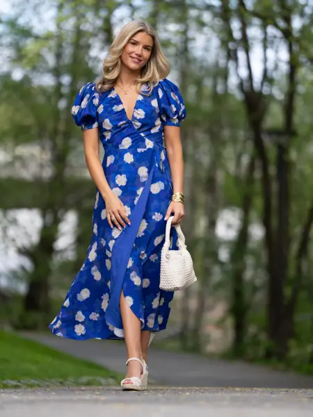 Ung jente i lang blå blomstrete kjole, sandaler og hvit håndveske