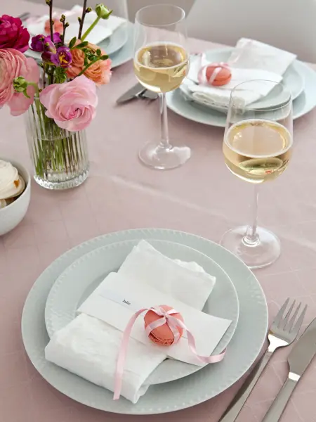 Dekket bord rosa duk, hvite servietter med rosa makron og navnelapp på, champagneglass og fargerike blomster