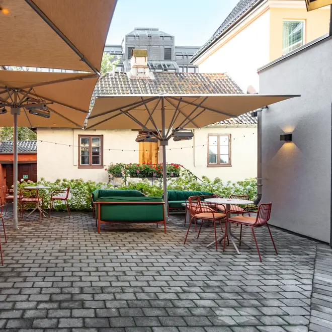Bakgård med brostein, røde runde bord med stoler rundt, og store parasoller over sitteplassene.