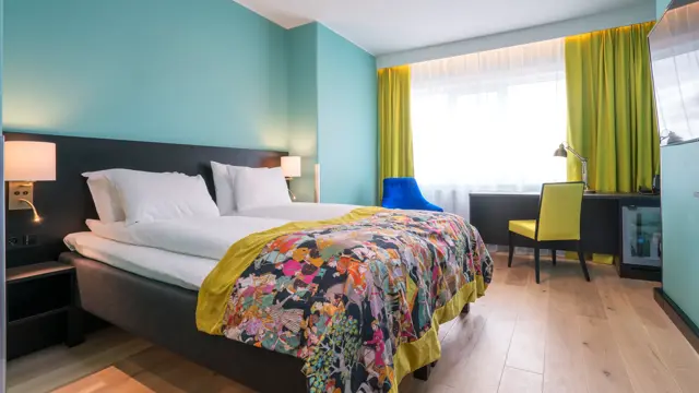 Twin room med hvit sengetøy, mønstret sengeteppe, gule gardiner, skrivepult og blå lenestol på Thon Hotel Europa i Oslo sentrum rett ved Slottsparken