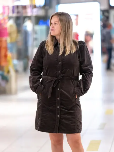 Jente med blondt hår, har på seg en knelang brun kjole med knapper og knytebelte i livet.