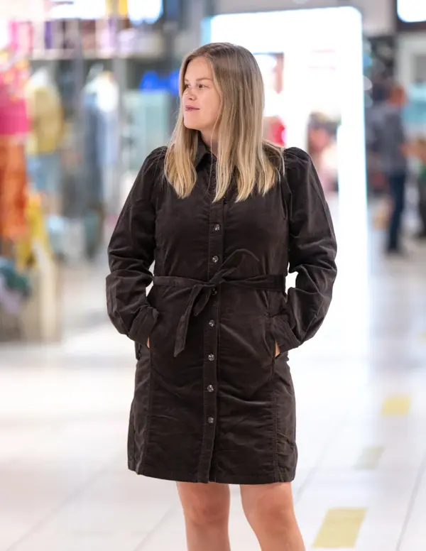 Jente med blondt hår, har på seg en knelang brun kjole med knapper og knytebelte i livet.