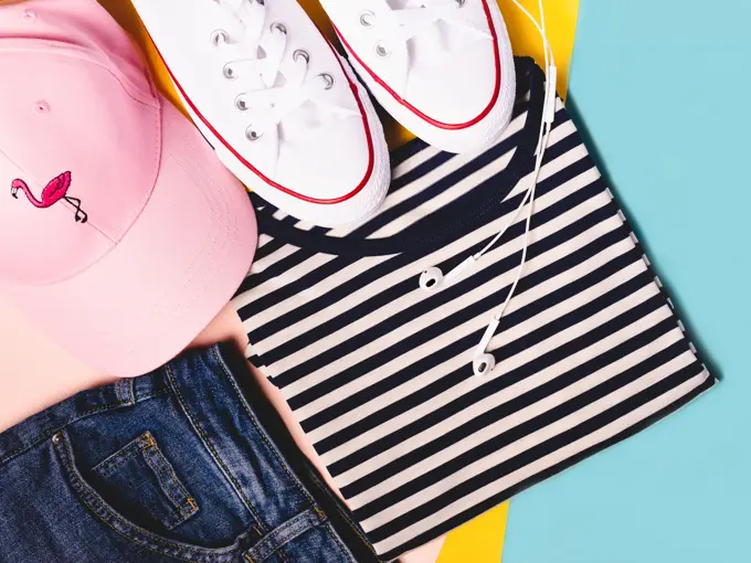 Fashion blogger concept. Summer sport, recreation and travel outfit: striped t-shirt, denim shorts, pink hat, white snickers. Minimal set of feminine teen clothes on pastel background