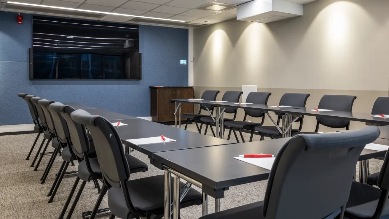 Møterom med u-bords oppsett, en vegg med blå tapet og TV skjerm på veggen. Penn og papir på bordet.