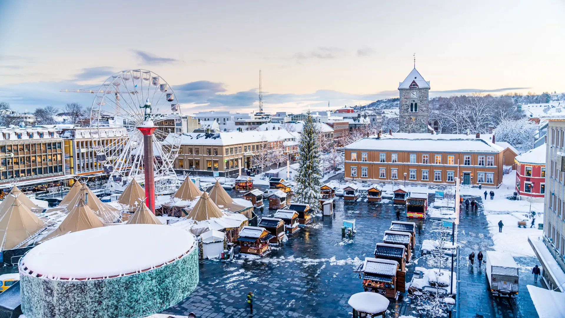 Julemarkedet på Torvet i Trondheim sett ovenfra, med pariserhjul, juletre, salgsboder og lavvoer i snødekt vinterlys.