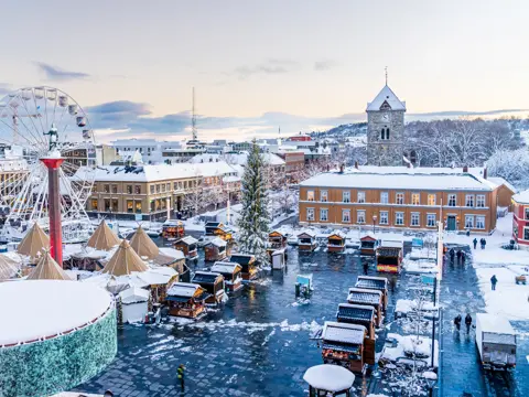 Julemarkedet på Torvet i Trondheim sett ovenfra, med pariserhjul, juletre, salgsboder og lavvoer i snødekt vinterlys.