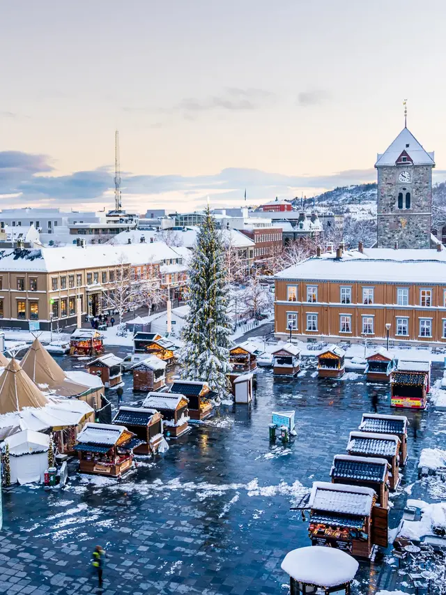 Julemarkedet på Torvet i Trondheim sett ovenfra, med pariserhjul, juletre, salgsboder og lavvoer i snødekt vinterlys.