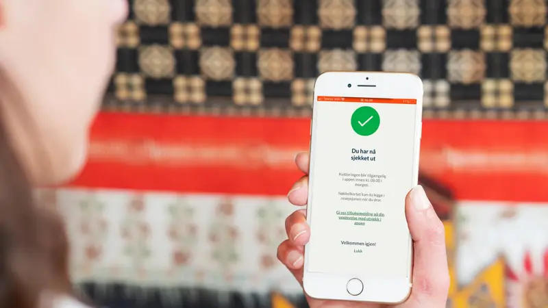 Dame som holder mobil oppe, på skjermen er det selvbetjent utsjekk