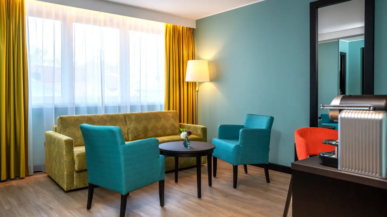 Sittegruppe med gul sofa, turkise stoler og sort rundt bord. Stort vindu med gule gardiner, turkis vegg med vegghengt speil på junior suite på Thon Hotel Maritim
