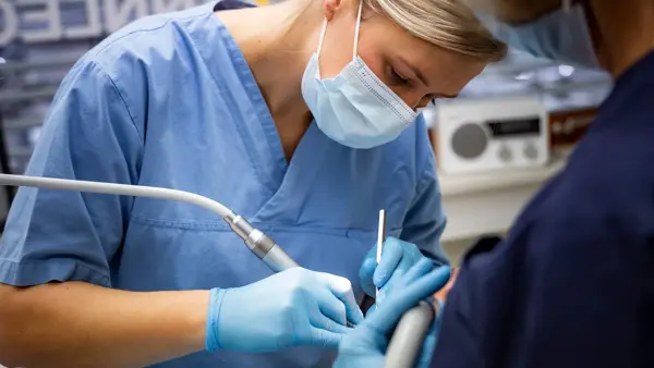 tannlege med munnbind, blå uniform og hansker som jobber med en kunde