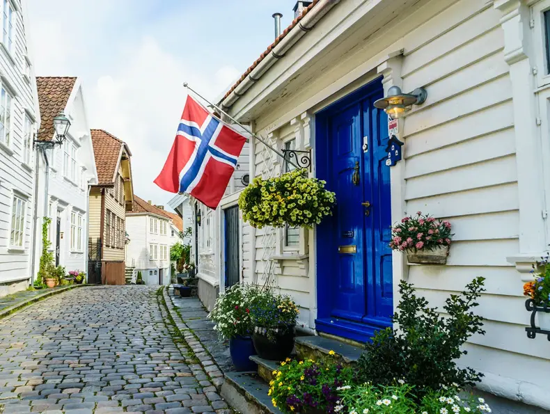 Brosteinsbelagt gate med hvite trehus, blå inngangsdør med flagg og grønne planter i forgrunn. Foto til artikkel om dekorasjoner til 17. mai.