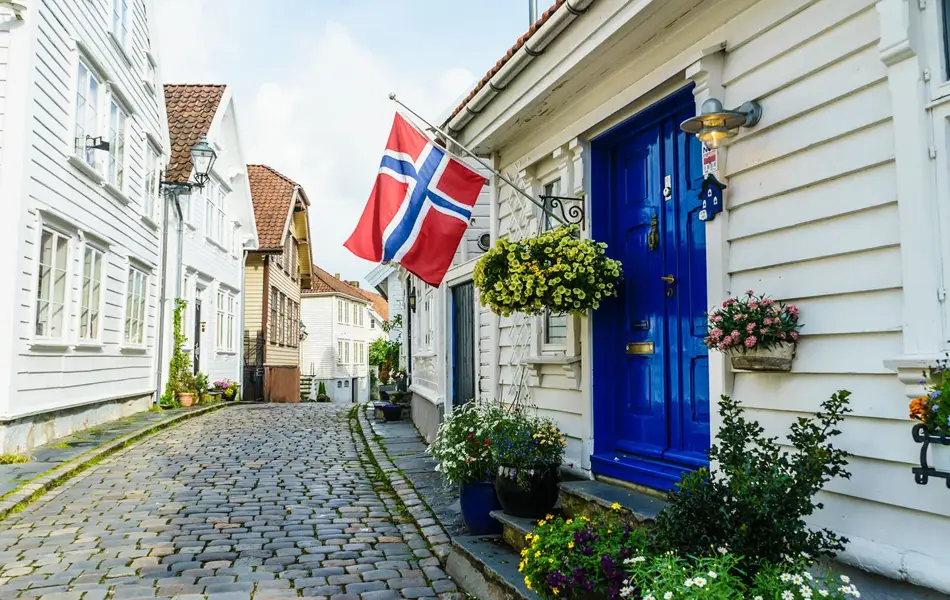 Brosteinsbelagt gate med hvite trehus, blå inngangsdør med flagg og grønne planter i forgrunn. Foto til artikkel om dekorasjoner til 17. mai.