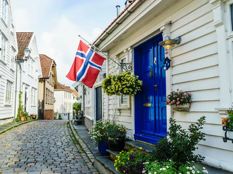 Brosteinsbelagt gate med hvite trehus, blå inngangsdør med flagg og grønne planter i forgrunn. Foto til artikkel om dekorasjoner til 17. mai.