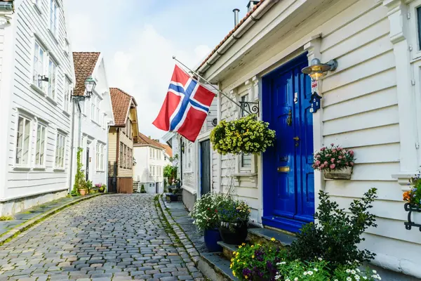 Brosteinsbelagt gate med hvite trehus, blå inngangsdør med flagg og grønne planter i forgrunn. Foto til artikkel om dekorasjoner til 17. mai.