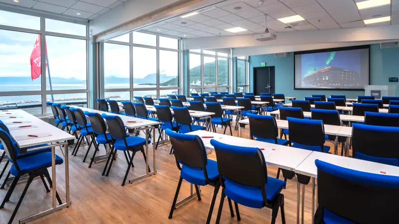 Møterom Vågsfjord, flere små skrivebord med blå stoler, 11 store vinduer fra gulv til tak, en projektor, parkettgulv med turkise vegger. 