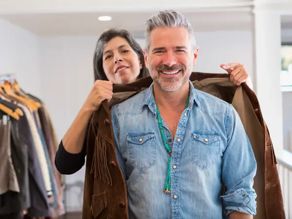 Mann i butikk iført denimskjorte som får hjelp til å prøve en western-inspirert brun semsket skinnjakke med frynser