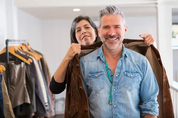 Mann i butikk iført denimskjorte som får hjelp til å prøve en western-inspirert brun semsket skinnjakke med frynser