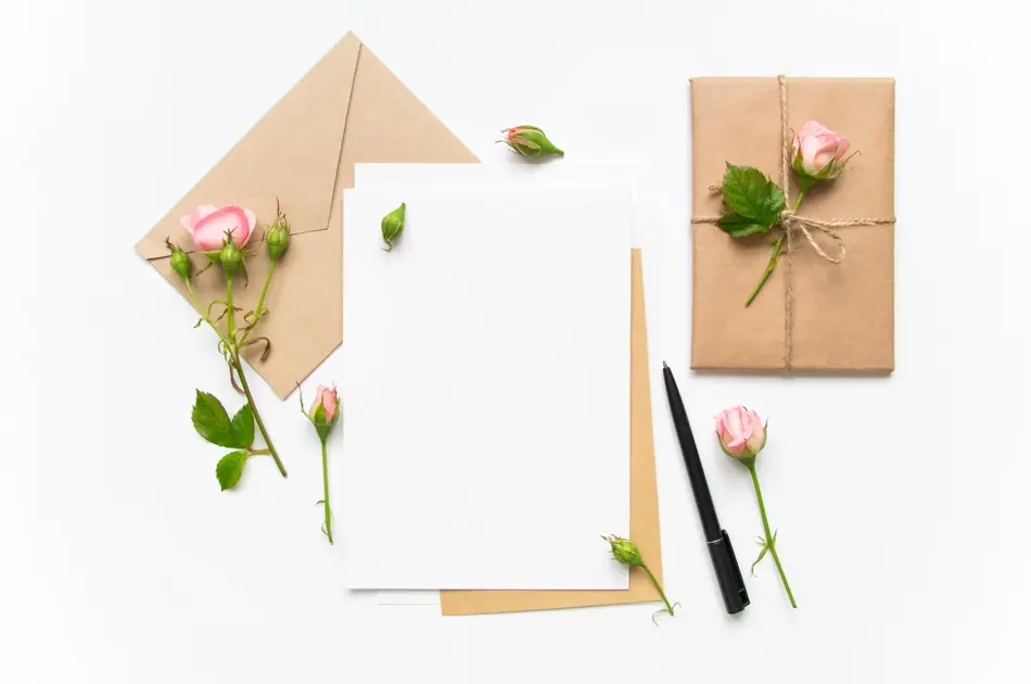 Letter, envelope and a present in eco paper on white background. Wedding invitation cards or love letter with pink roses. Valentine's day or other holiday concept, top view, flat lay, overhead view