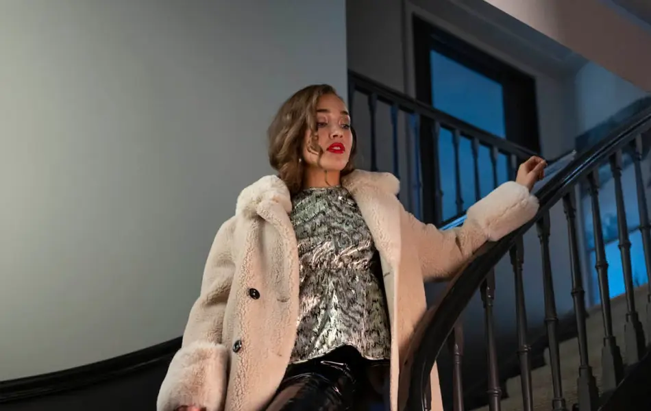 Damemodell står i trappen med lakkbukser, lakkboots, sølvglitrende topp og hvit teddykåpe. Holder en sort veske.