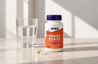 En boks med Vitamin D3 og K2 fra Now står ved siden av et glass med vann.