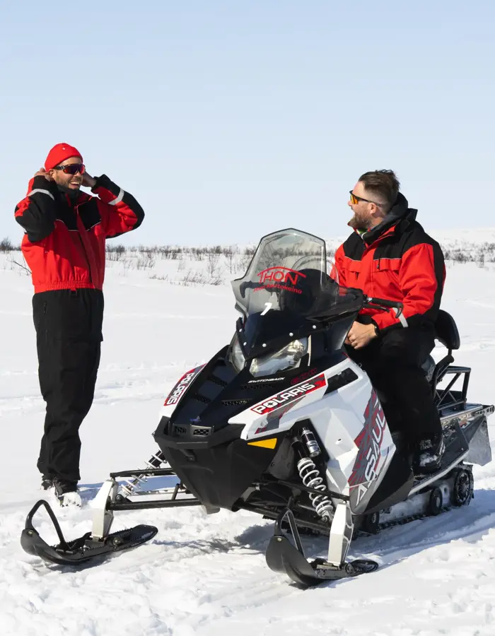 En person sitter på snøscooter og snakker med en person som står ved siden av, i snødekt landskap.