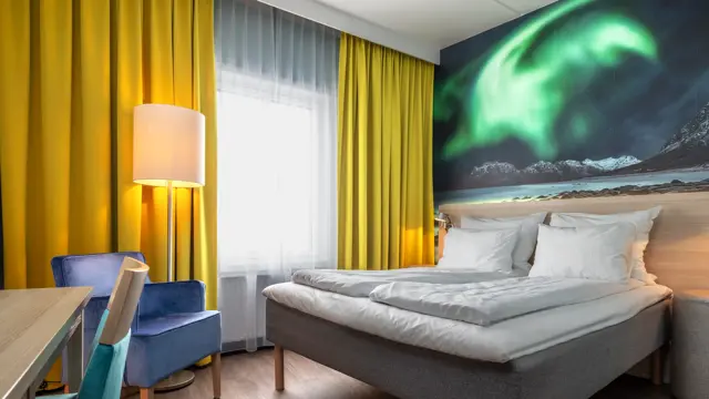 Dobbeltrom med dobbeltseng, trehvit sengegavl, stort nordlys bilde på veggen, gule gardiner, lilla lenestol, skrivebord med turkis stol.