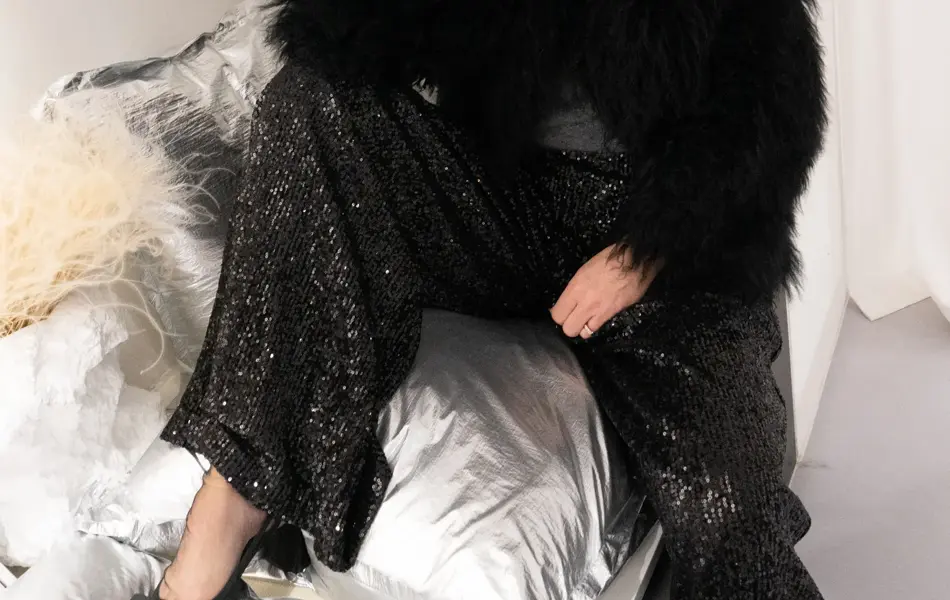 Dame med sort fuskepelsjakke, sølvtopp, glitrende bukser og pumps med sløyfe.