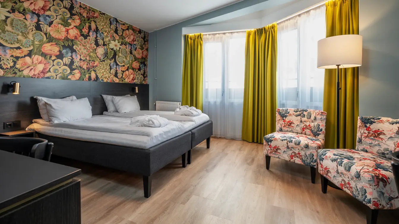 Business rom med dobbeltseng, to mønstrede lenestoler, gule gardiner og fargerik tapet på vegg bak seng.