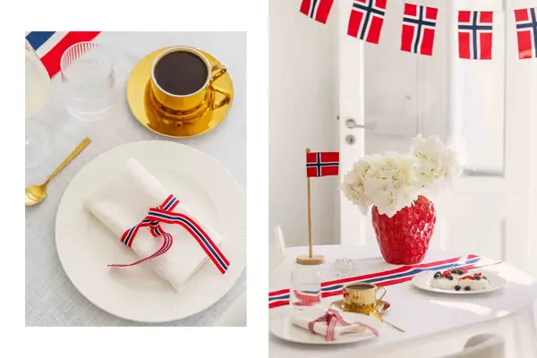 Hvit tallerken med hvit serviett og bånd bundet runt, gull skjer og kaffekopp, flagg lenge, bord med hvit duk, rød vase med hvite blomster, bordflagg.