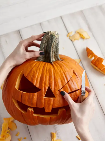 Woman carving big orange pumpkin into jack-o-lantern for Halloween holiday decoration on white wooden planks, close up view