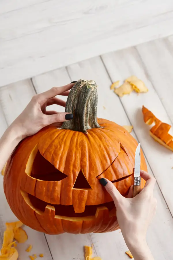 Woman carving big orange pumpkin into jack-o-lantern for Halloween holiday decoration on white wooden planks, close up view