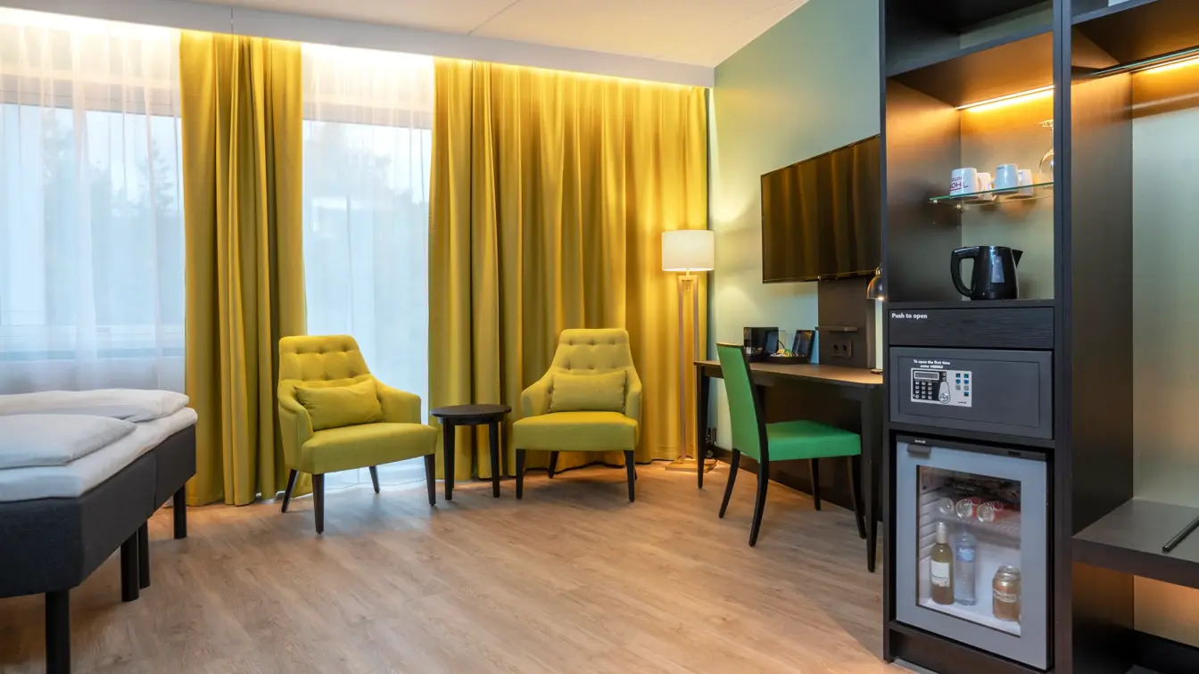 Sittegruppe med gule stoler, gule gardiner, skrivebord med grønn stol, TV, minibar og vannkoker i business rom på Thon Hotel Linne