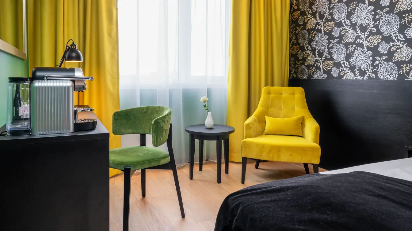 Mørk mønstrete tapet med blomster, gul stol med rundt bord, gule gardiner, skrivebord med kaffemaskin og grønn stol i superior rom på Thon Hotel Kirkenes.