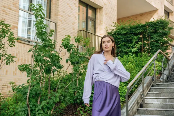 Kvinne står ute i en trapp foran grønne vekster iført lilla skjørt og dus lilla bluse. 